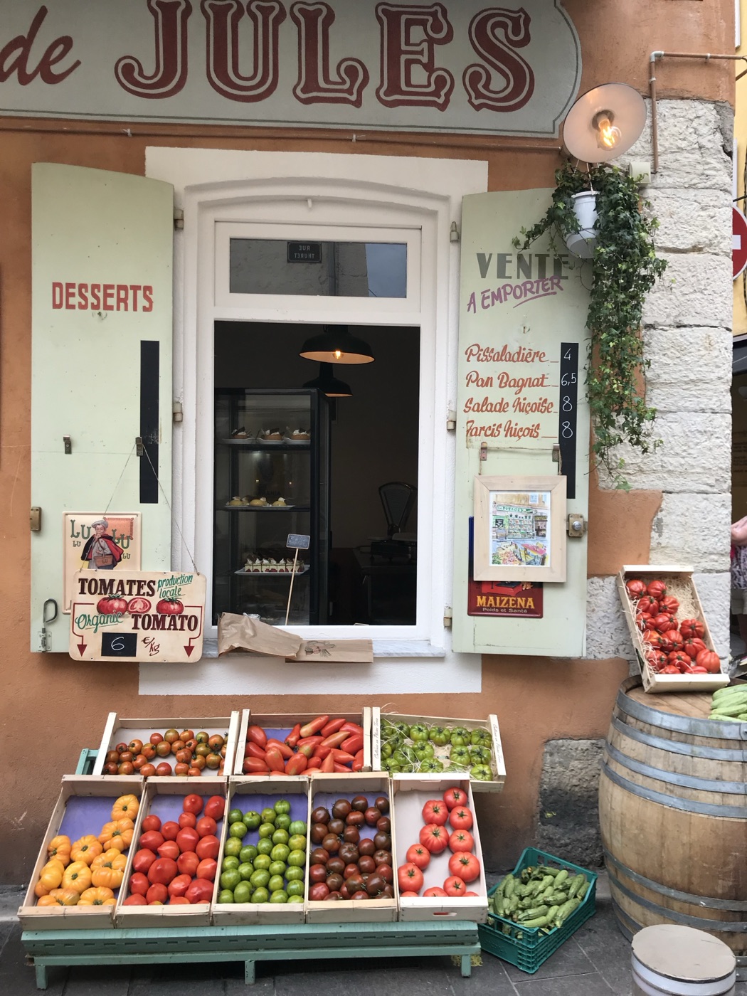 A sunlit French market stall named 'de Jules', its crates piled with fresh tomatoes, apples and oranges beside wooden wine barrels